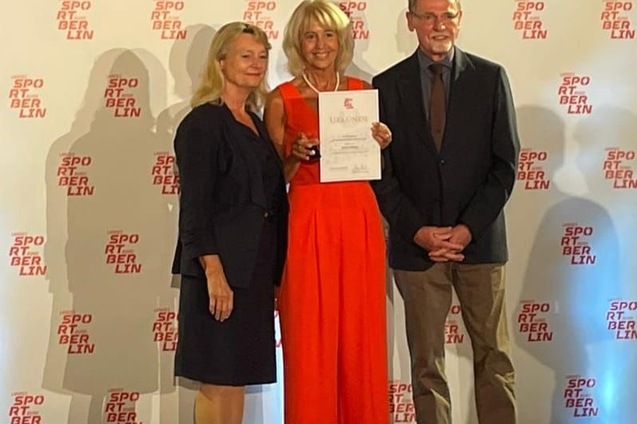 Drei Personen stehen vor einem Hintergrund mit dem Logo "Sport Berlin" und halten eine Urkunde in der Hand.