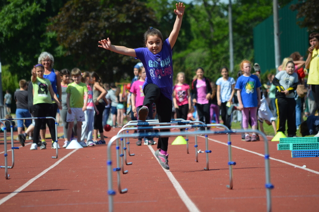 M&auml;dchen in lila T-Shirt springt &uuml;ber H&uuml;rden auf einem Sportplatz, umgeben von anderen Kindern und Zuschauern.