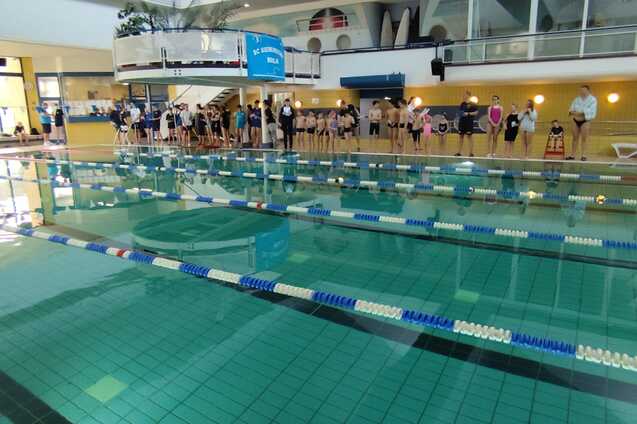 Schwimmbecken mit mehreren Bahnen und zahlreichen wartenden Schwimmern am Rand des Pools in einem Hallenbad.
