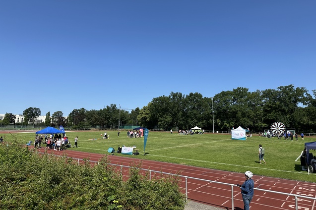 Sportveranstaltung auf einem gr&uuml;nen Feld mit St&auml;nden, Besuchern und einem Gl&uuml;cksrad vor klarem Himmel.