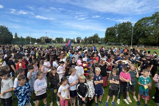 Gro&szlig;e Gruppe von Kindern in sportlicher Kleidung, versammelt auf einem Rasensportplatz unter blauem Himmel.