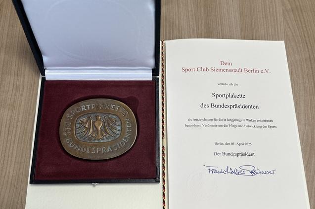 Medaille der Sportplakette des Bundespr&auml;sidenten in der Box, daneben ein Urkundendokument auf einem Tisch.
