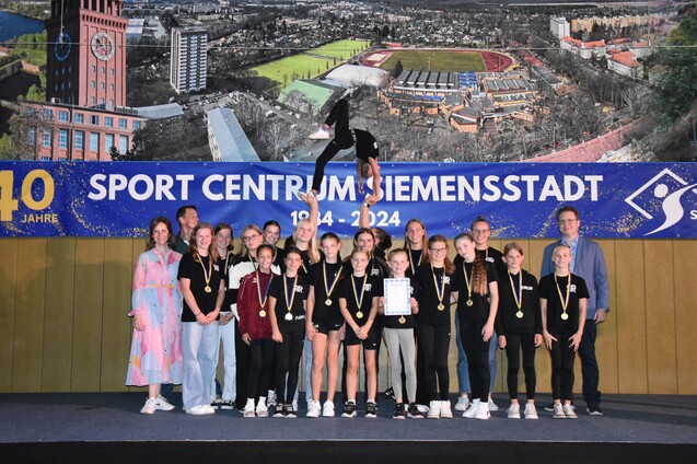 Gruppe von Sportlern mit Medaillen auf B&uuml;hne, w&auml;hrend eine Akrobatin eine Darbietung zeigt, im Hintergrund ein gro&szlig;es Banner.