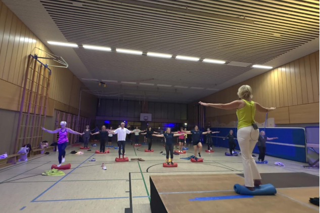Gruppentraining in einer Sporthalle: Teilnehmer stehen auf Balance-Kissen und folgen einer Trainerin mit erhobenen Armen.