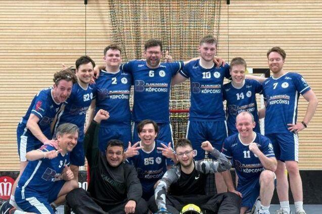 Gruppennfoto einer Handballmannschaft in blauen Trikots, l&auml;chelnd und posierend in einer Sporthalle.