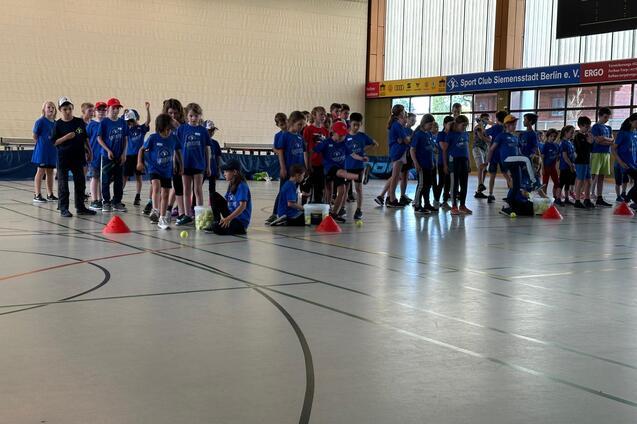 Gruppierung von Kindern in blauen T-Shirts in einer Sporthalle, angereiht an roten Kegeln mit Tennisb&auml;llen.