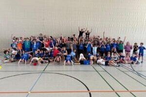 Gruppenfoto von &uuml;ber 70 Kindern in blauen T-Shirts, die freudig in einer Sporthalle posieren.