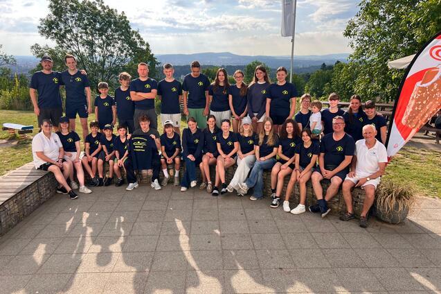 Gruppenfoto von 24 Personen in blauen T-Shirts, im Freien mit Bäumen und Blick auf die Landschaft.