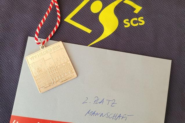 Medaille mit rot-weißer Schnur, Umschlag mit Aufschrift „2. Platz Mannschaft“ auf einem dunklen Trikot.