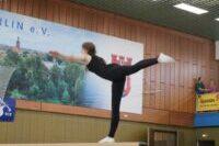 Turnerin in schwarzem Outfit balanciert auf einem Schwebebalken in einer Turnhalle. Zuschauer im Hintergrund.