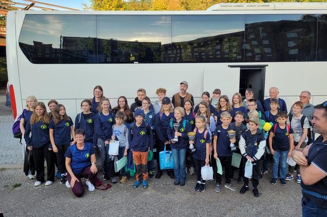 Gruppe von Kindern und Erwachsenen in Sportkleidung vor einem Reisebus, einige halten Pokale in den H&auml;nden.