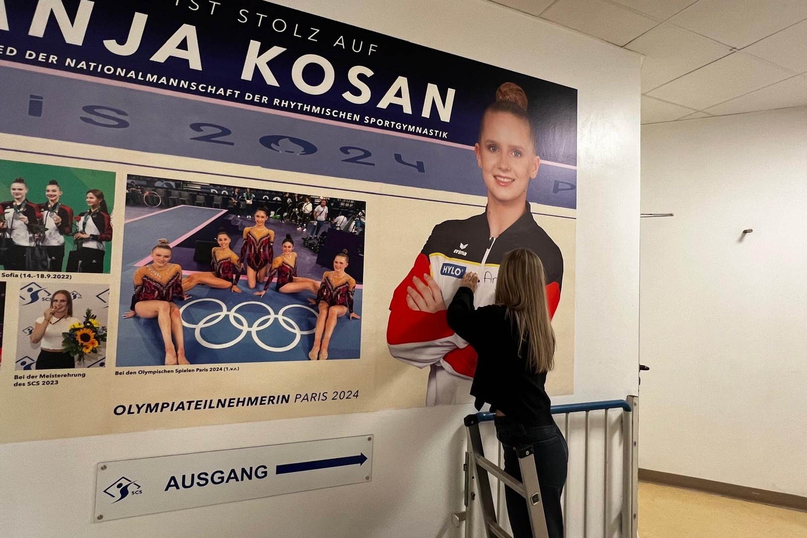 Frau auf einer Leiter, die ein Bild von Anja Kosan, einer Sportlerin, an einer Wand anbringt. Olympische Ringe sichtbar.