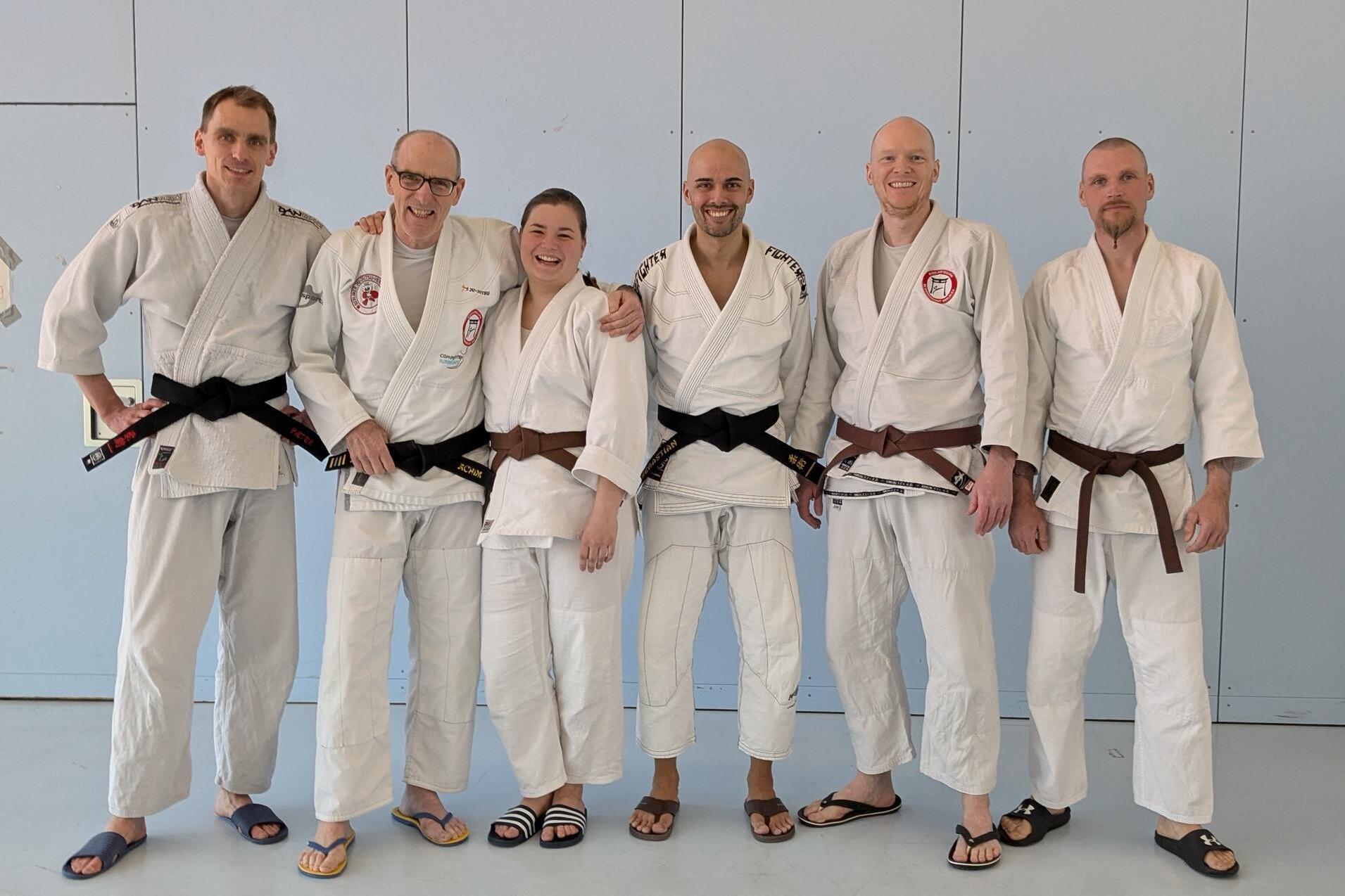 Gruppe von f&uuml;nf Judokas in Judogis, zwei mit braunen und drei mit schwarzen G&uuml;rteln, stehen l&auml;chelnd in einer Trainingshalle.