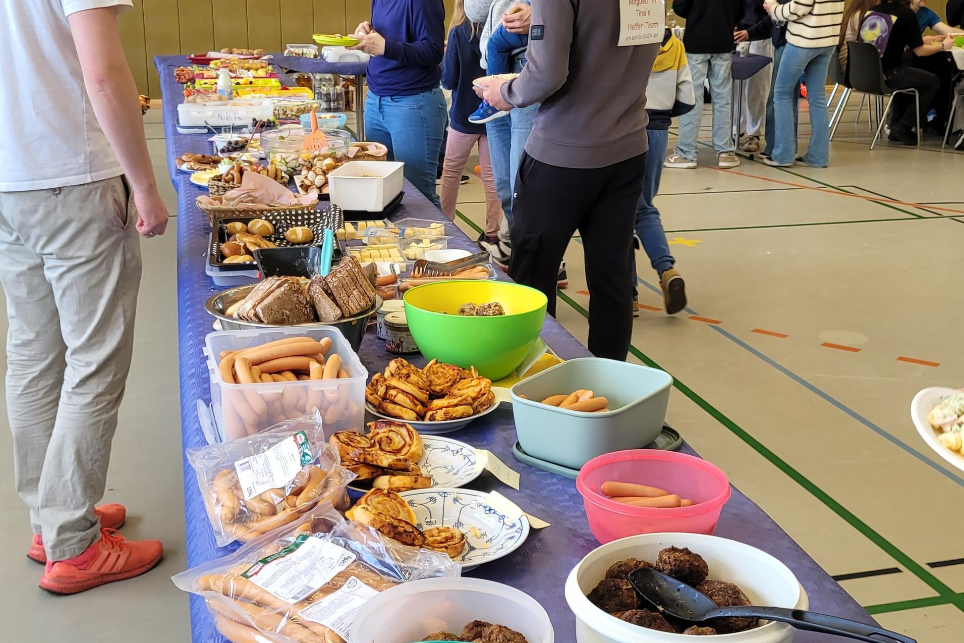 Buffet mit verschiedenen Speisen, unter anderem Salate, Snacks und Wurst, w&auml;hrend G&auml;ste sich unterhalten.