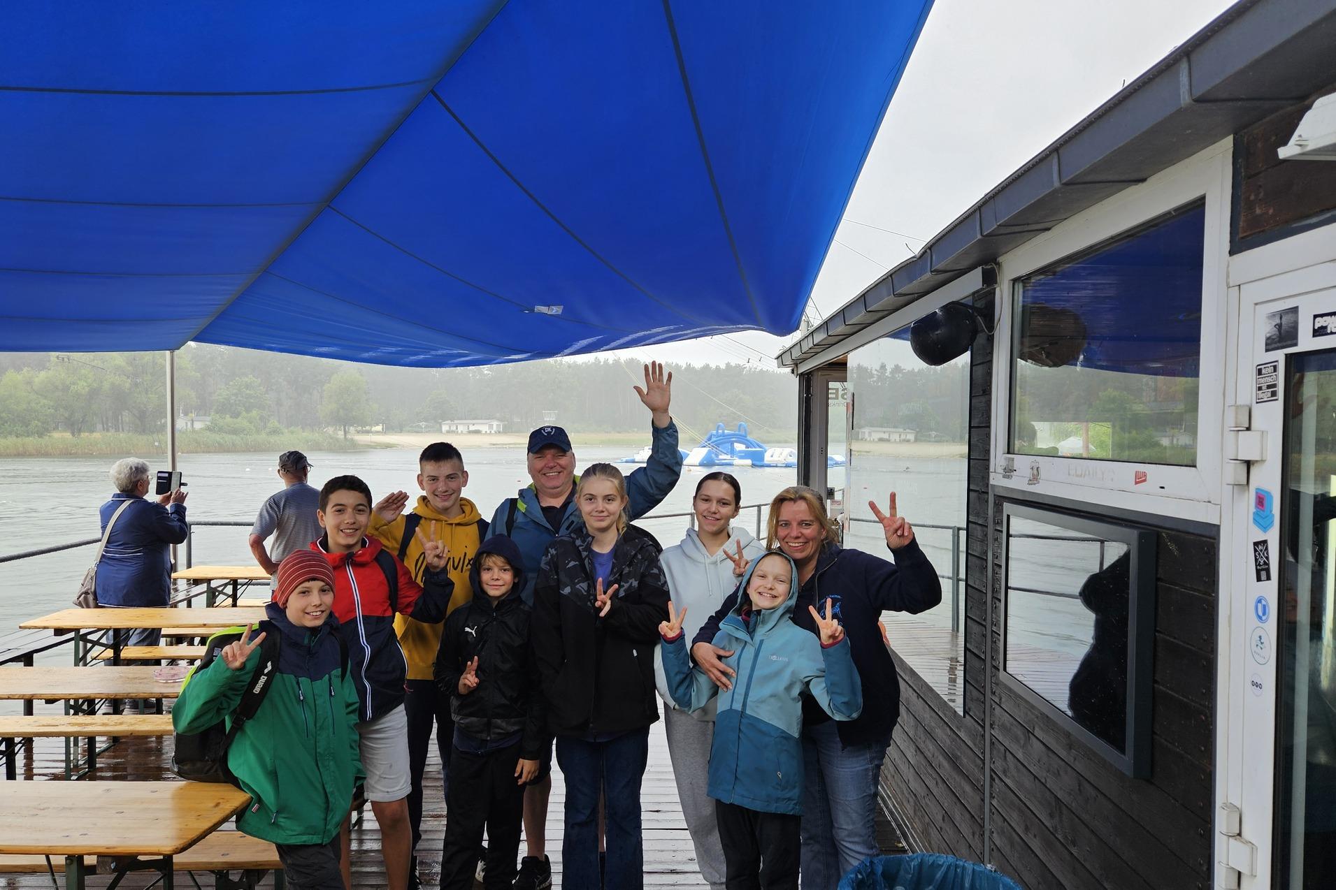 Gruppe von acht Personen steht unter einem blauen Pavillon an einem Gew&auml;sser, einige zeigen Victory-Zeichen.