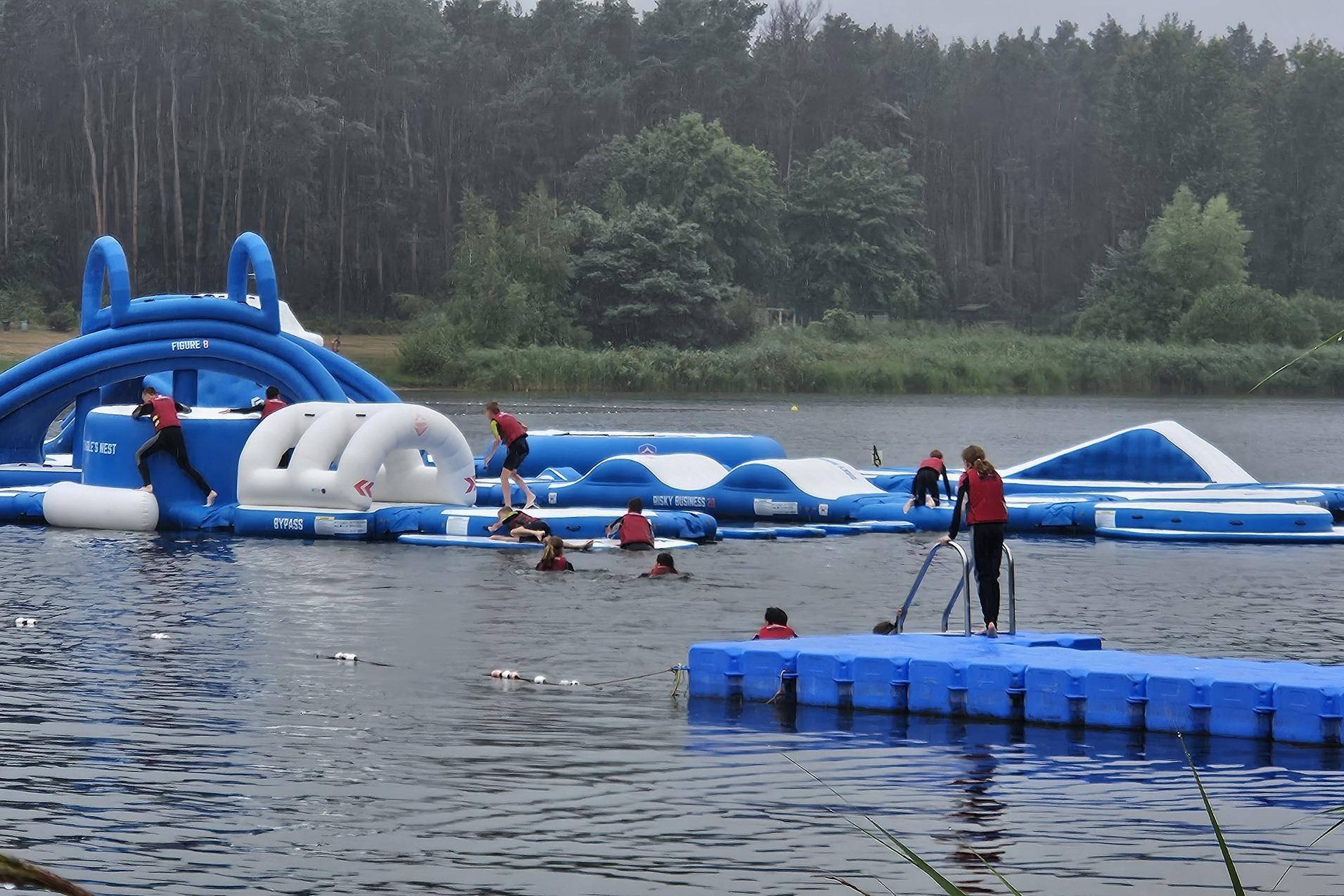 Gruppe von Personen auf einem schwimmenden Hindernisparkour im Wasser, umgeben von B&auml;umen und einem grauen Himmel.