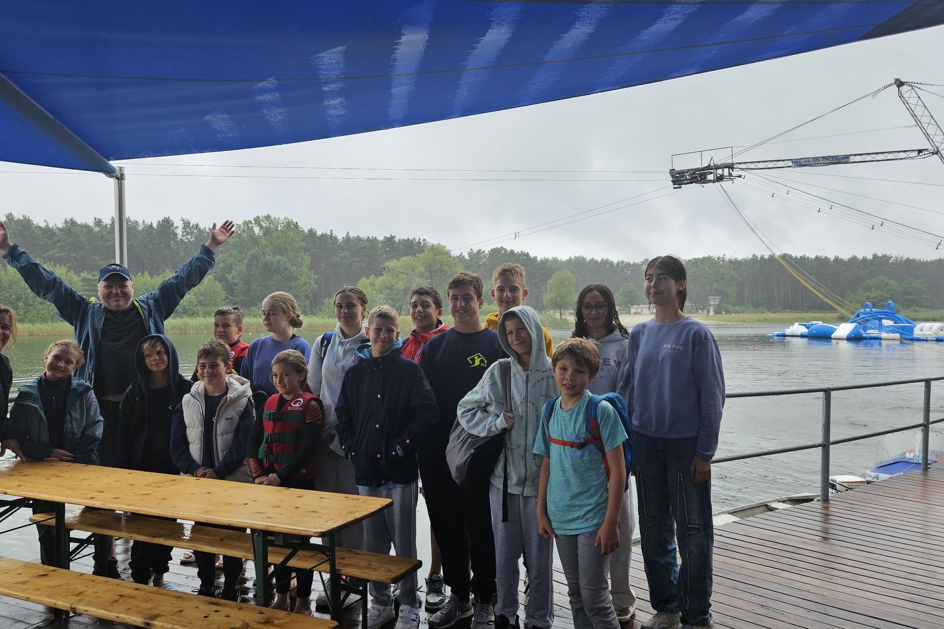 Gruppe von Jugendlichen und Erwachsenen unter einem blauen Sonnenschutz am Wasser, mit Wassersportger&auml;ten im Hintergrund.