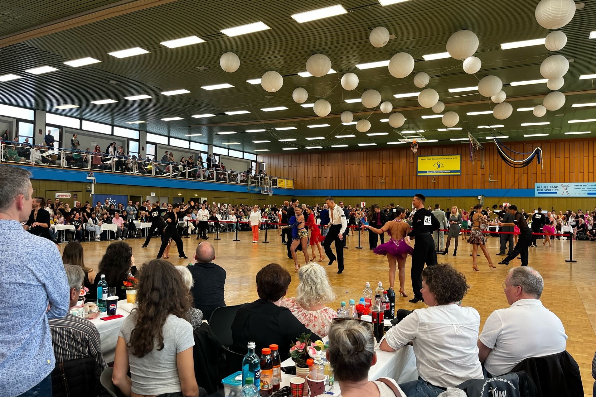 Tanzpaare f&uuml;hren synchron T&auml;nze auf einem Wettbewerb auf, Zuschauer betrachten das Geschehen in einer gro&szlig;en Halle.
