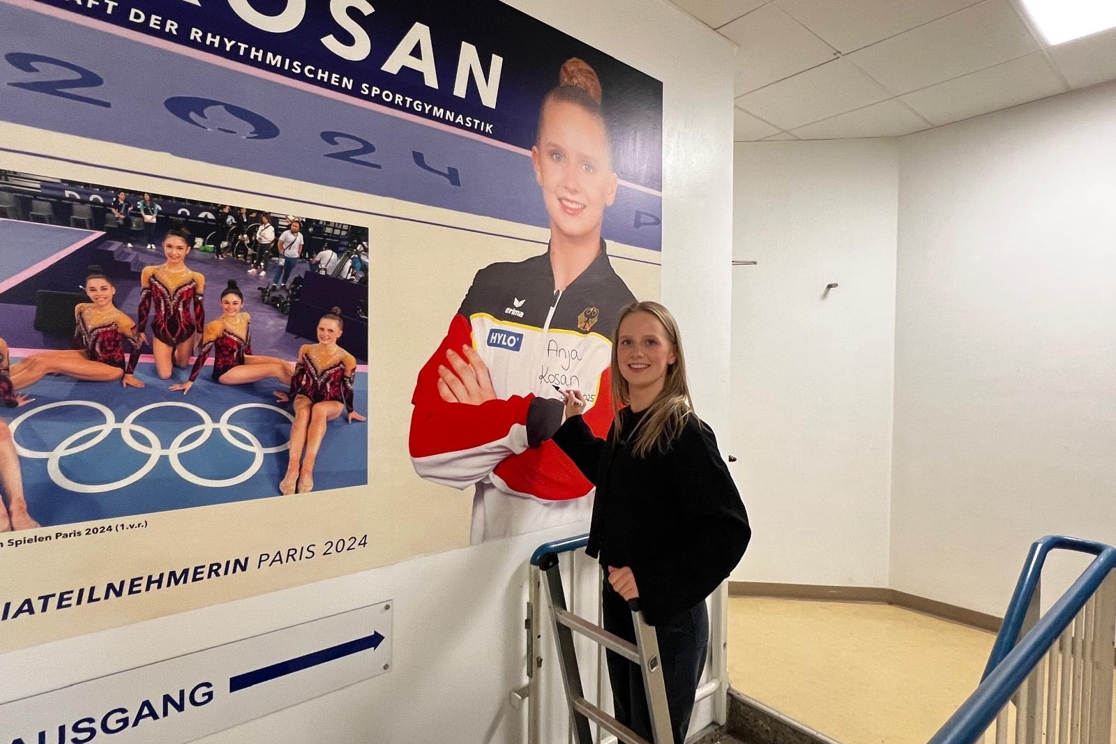 Junge Frau steht an einer Wand mit einem Plakat von Anja Kosan, bevor sie an den Olympischen Spielen 2024 teilnehmen.