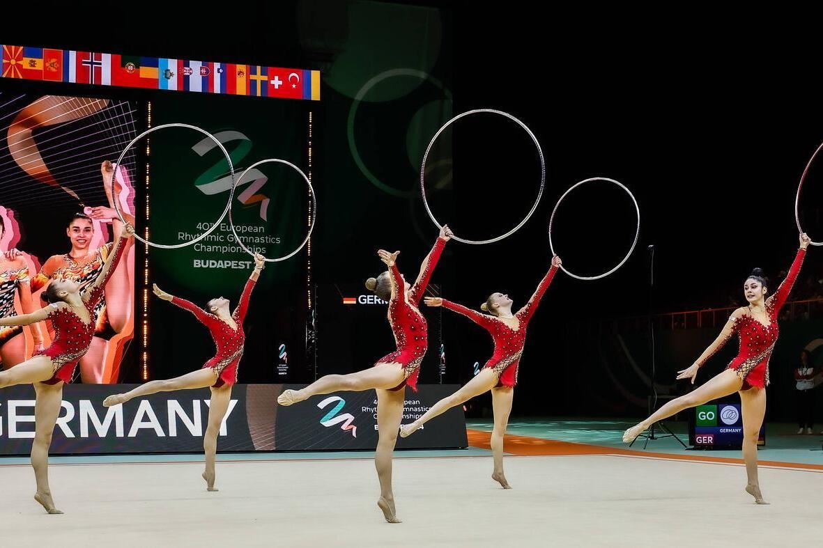 F&uuml;nf weibliche Turnerinnen in roten Kost&uuml;men synchronisieren Bewegungen mit Reifen auf einer Gymnastikfl&auml;che.