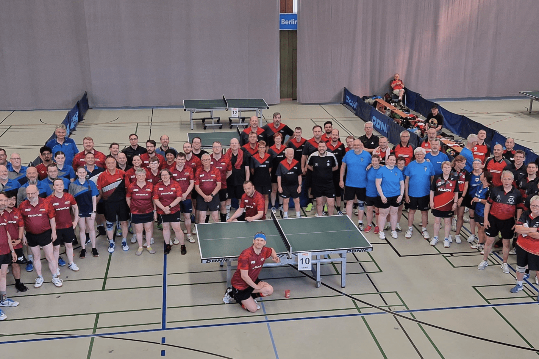 Gruppenfoto von Tischtennisspielern in Sportkleidung auf einer Sporthalle mit Tischtennisplatten im Hintergrund.