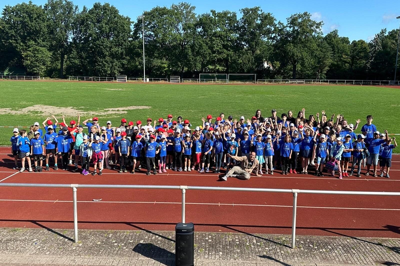 Gruppierung von Kindern in blauen T-Shirts und roten Kappen auf einer Laufbahn, umgeben von B&auml;umen und einem gr&uuml;nen Feld.