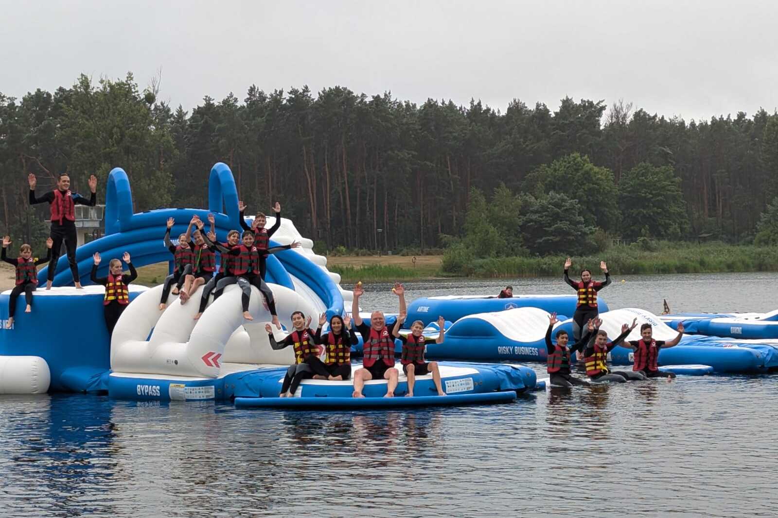 Gruppierung von Personen in Schwimmwesten, die auf schwimmenden Plattformen an einem See spielen und winken.