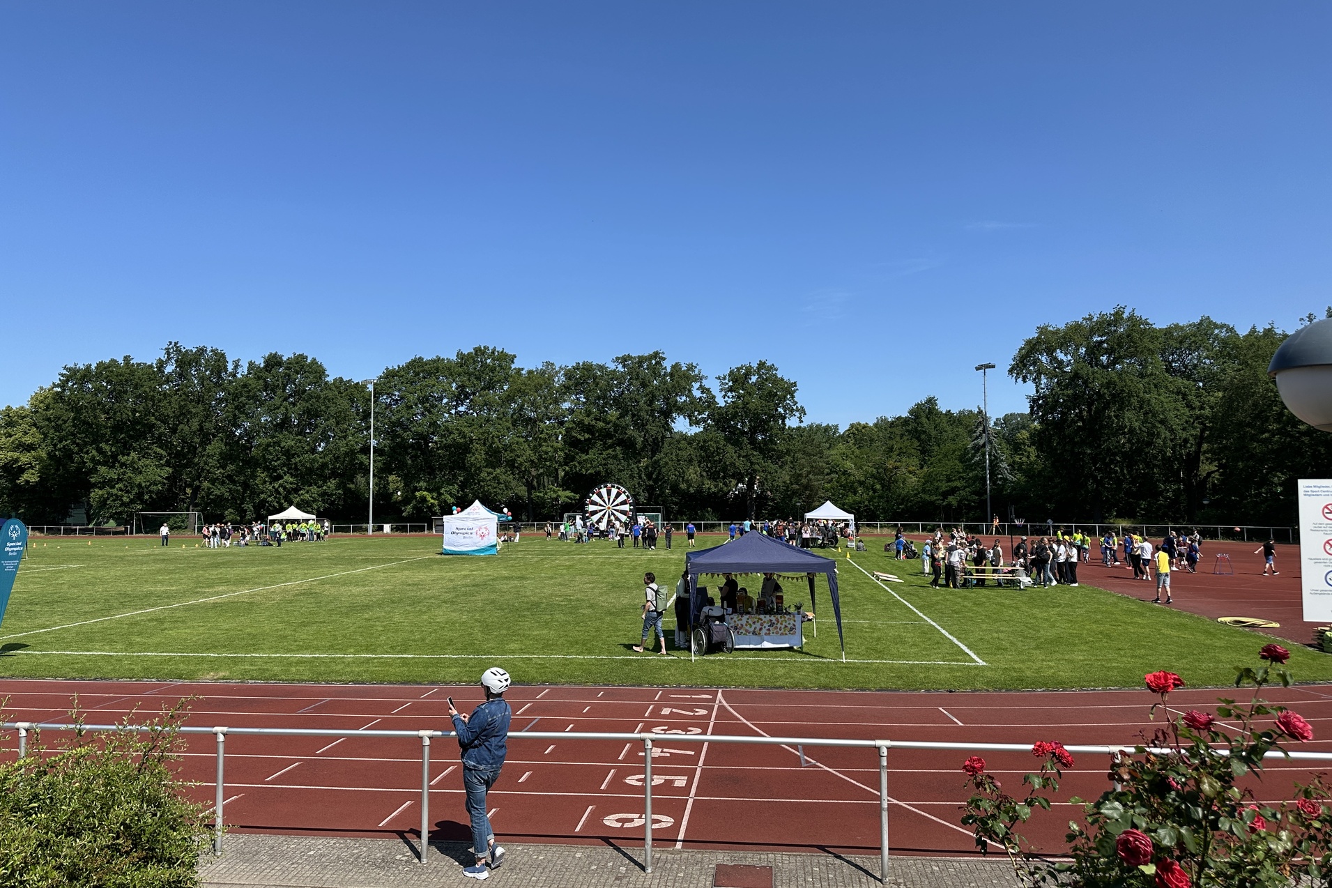 Veranstaltung auf einem Sportplatz mit St&auml;nden, Besuchern und einem gro&szlig;en Gl&uuml;cksrad unter blauem Himmel.