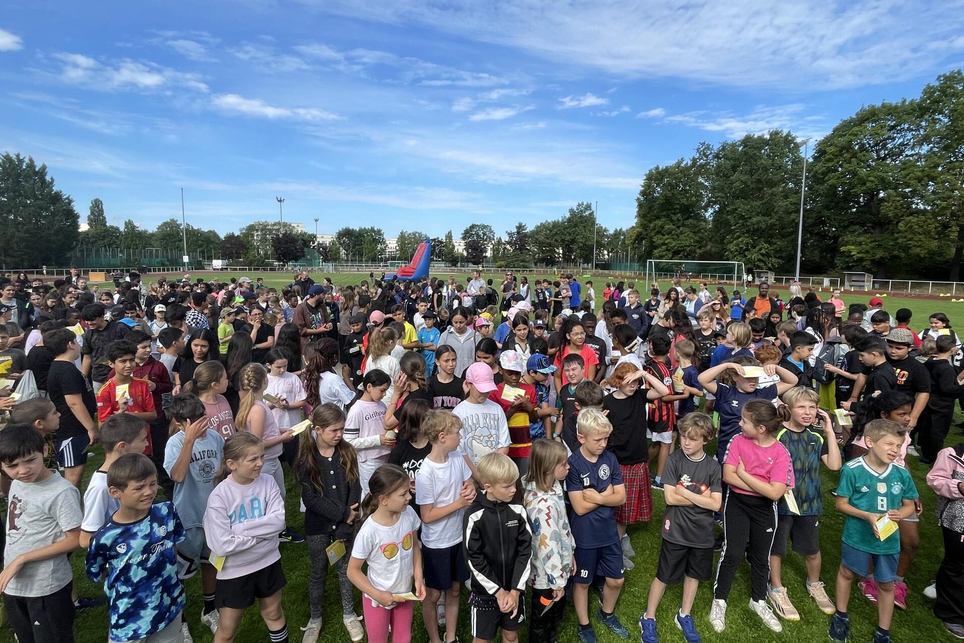 Gro&szlig;e Gruppe von Kindern in sportlicher Kleidung, versammelt auf einem Rasensportplatz unter blauem Himmel.