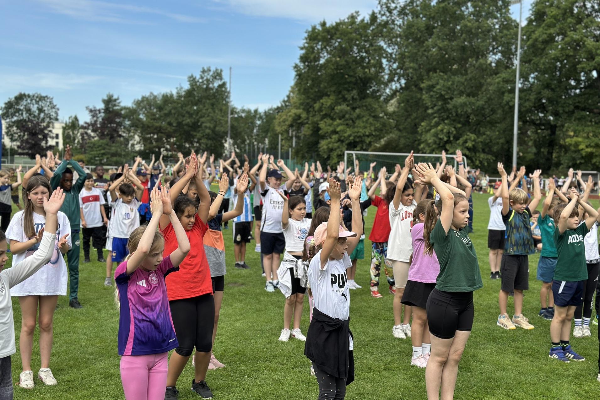 Gruppierung von Kindern in Sportkleidung, die im Freien mit erhobenen H&auml;nden &Uuml;bungen ausf&uuml;hren.