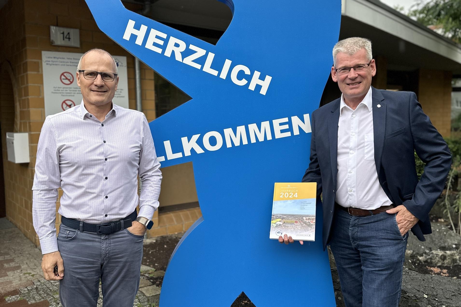 Zwei Männer posieren neben einem blauen Bären mit der Aufschrift „HERZLICH WILLKOMMEN“ vor einem Restaurant.