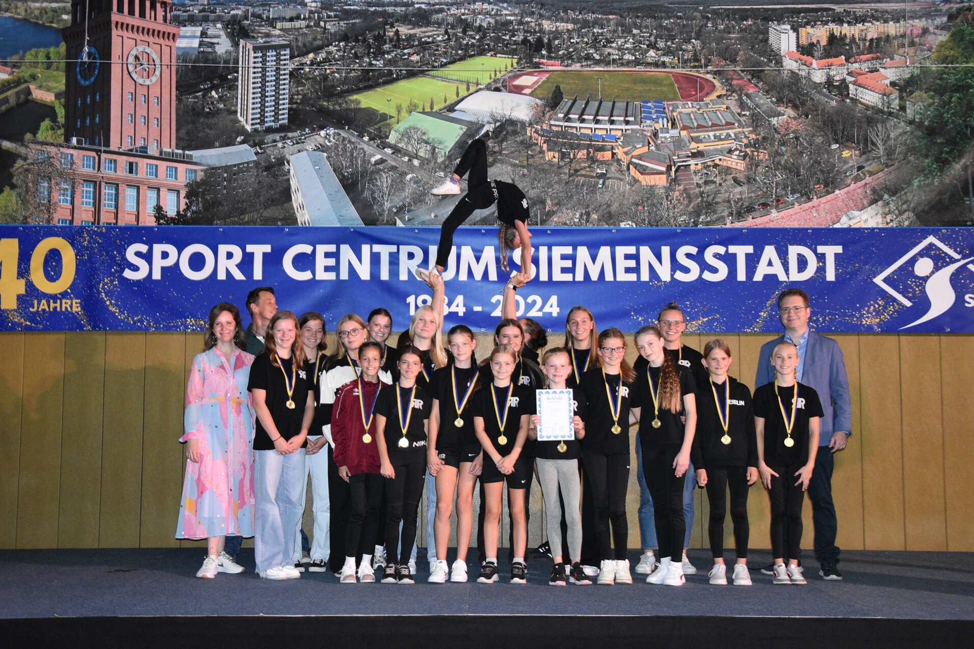 Gruppe von Sportlern mit Medaillen auf B&uuml;hne, w&auml;hrend eine Akrobatin eine Darbietung zeigt, im Hintergrund ein gro&szlig;es Banner.