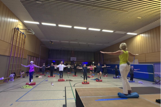 Gruppentraining in einer Sporthalle: Teilnehmer stehen auf Balance-Kissen und folgen einer Trainerin mit erhobenen Armen.