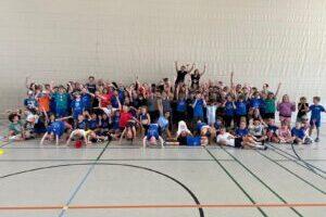 Gruppenfoto von &uuml;ber 70 Kindern in blauen T-Shirts, die freudig in einer Sporthalle posieren.