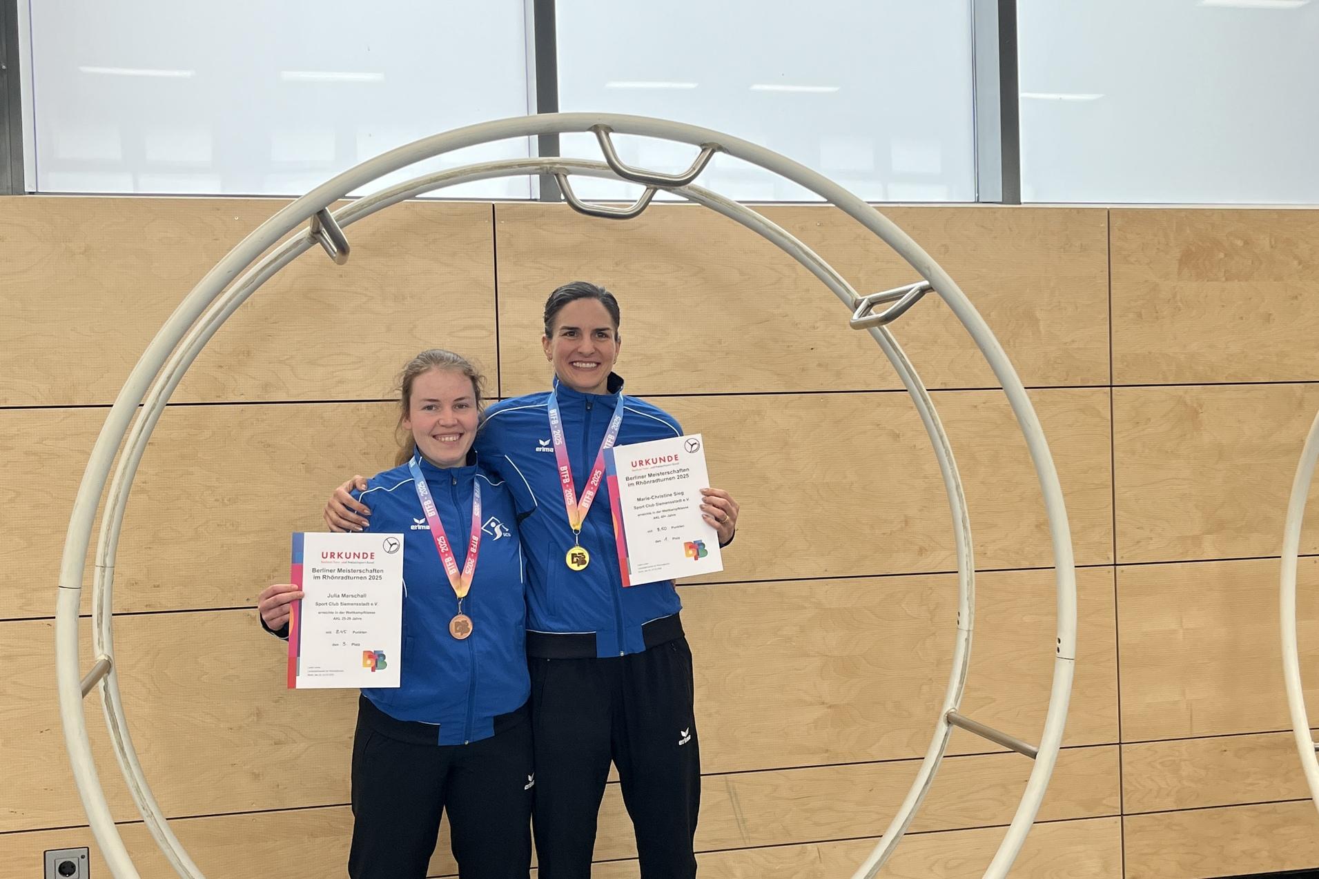 Zwei Athletinnen in blauen Sportjacken stehen in einem gro&szlig;en Holzring und halten Medaillen und Urkunden in den H&auml;nden.