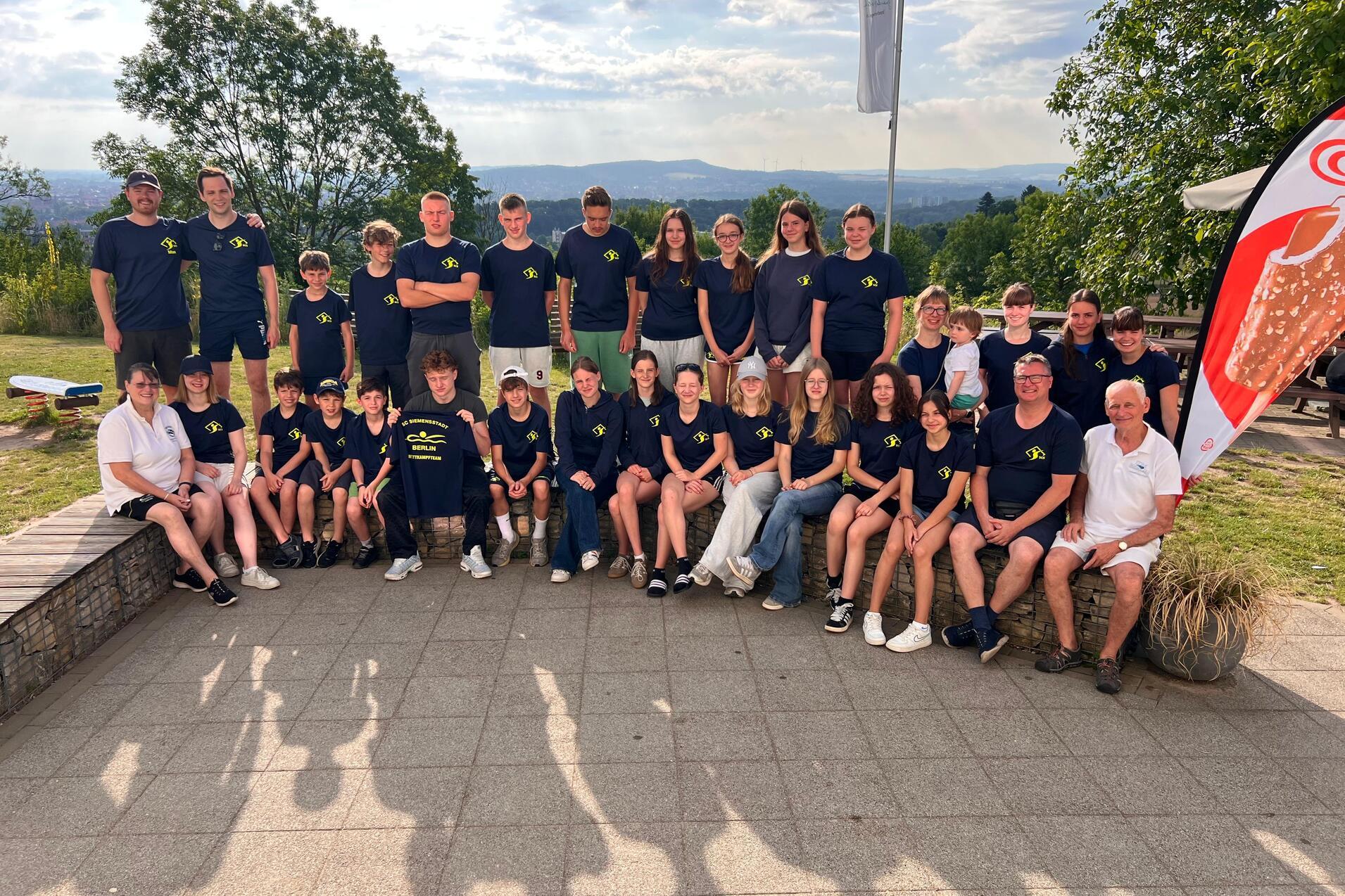 Gruppenfoto von 24 Personen in blauen T-Shirts, im Freien mit B&auml;umen und Blick auf die Landschaft.