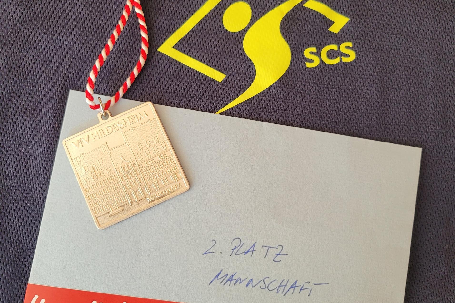 Medaille mit rot-wei&szlig;er Schnur, Umschlag mit Aufschrift &bdquo;2. Platz Mannschaft&ldquo; auf einem dunklen Trikot.