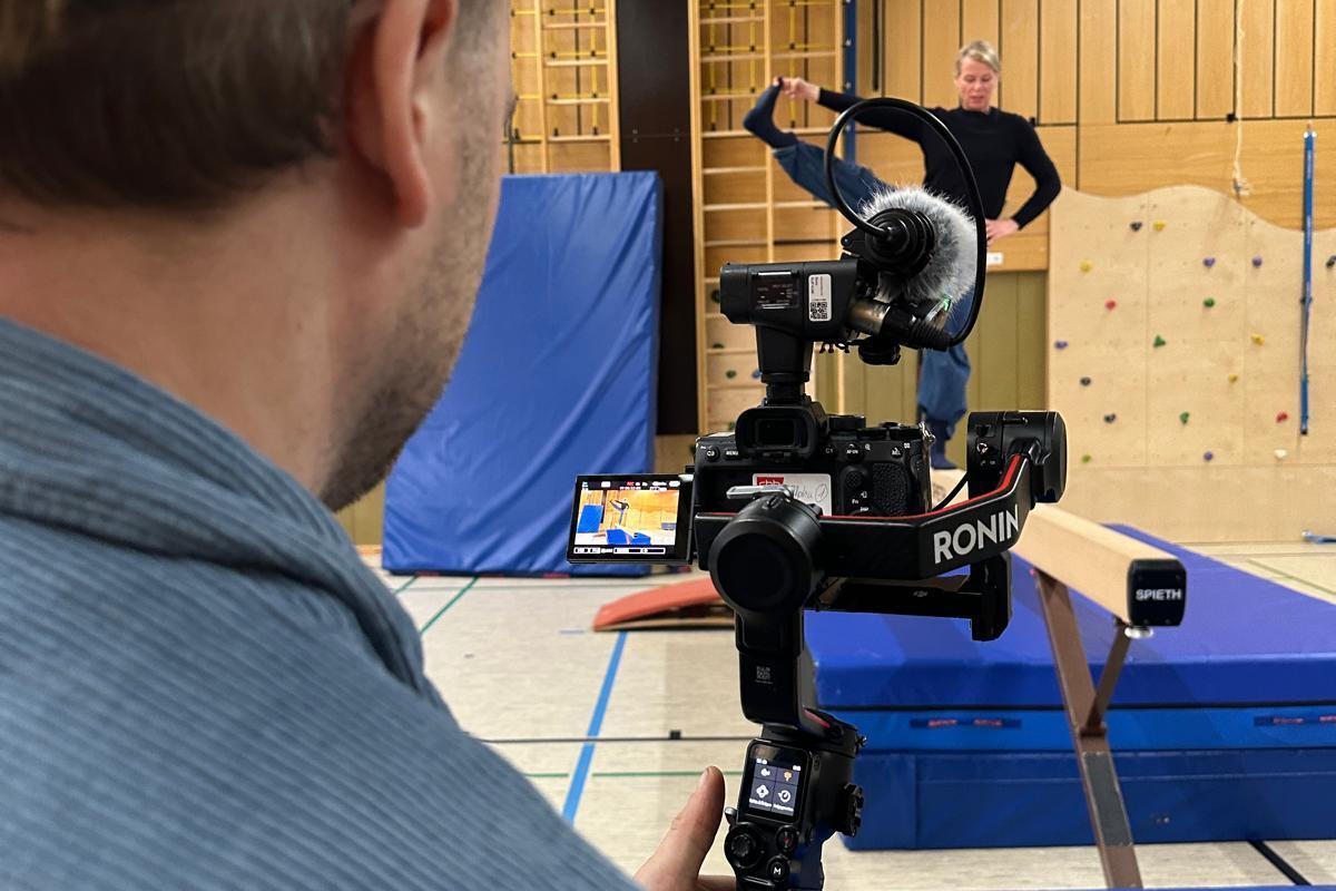 Person filmt eine Gymnastik&uuml;bung in einer Turnhalle mit Kamera auf einem Stabilisator und einem Balancebalken im Vordergrund.