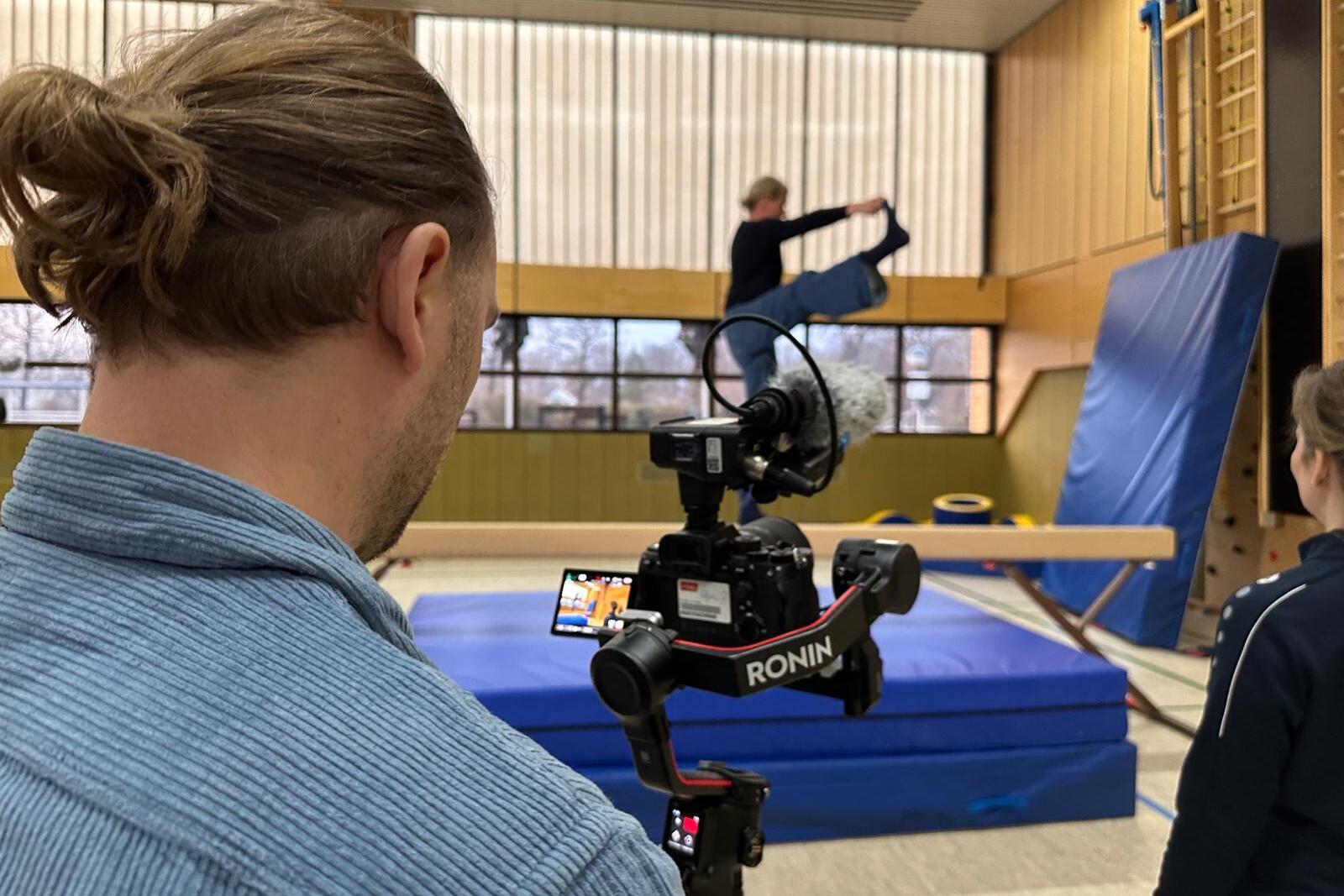 Eine Person filmt mit einer Kamera, w&auml;hrend eine andere Person eine &Uuml;bung an einem Schwebebalken demonstriert.