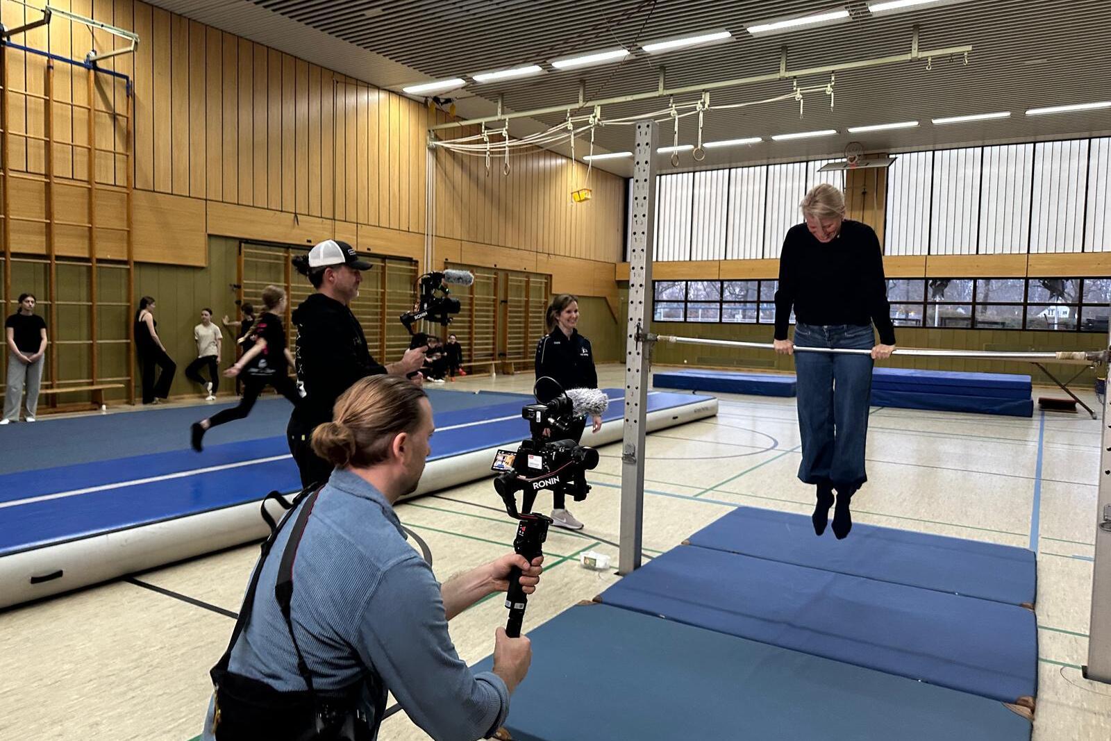 Person beim Training an einem Reck in einer Sporthalle, w&auml;hrend Videoaufnahmen durchgef&uuml;hrt werden.