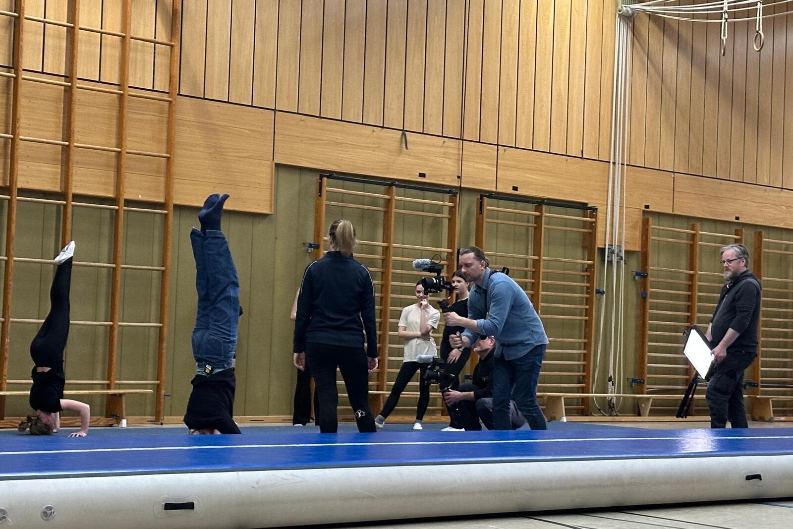 Gruppen&uuml;bung in einer Sporthalle: Zwei Personen machen Handst&auml;nde, w&auml;hrend mehrere Zuschauer das Geschehen beobachten.