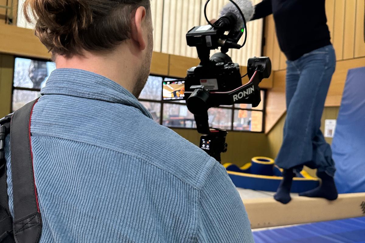 Ein Mann filmt eine Frau, die auf einem Balken in einer Turnhalle balanciert, w&auml;hrend er eine Kamera h&auml;lt.