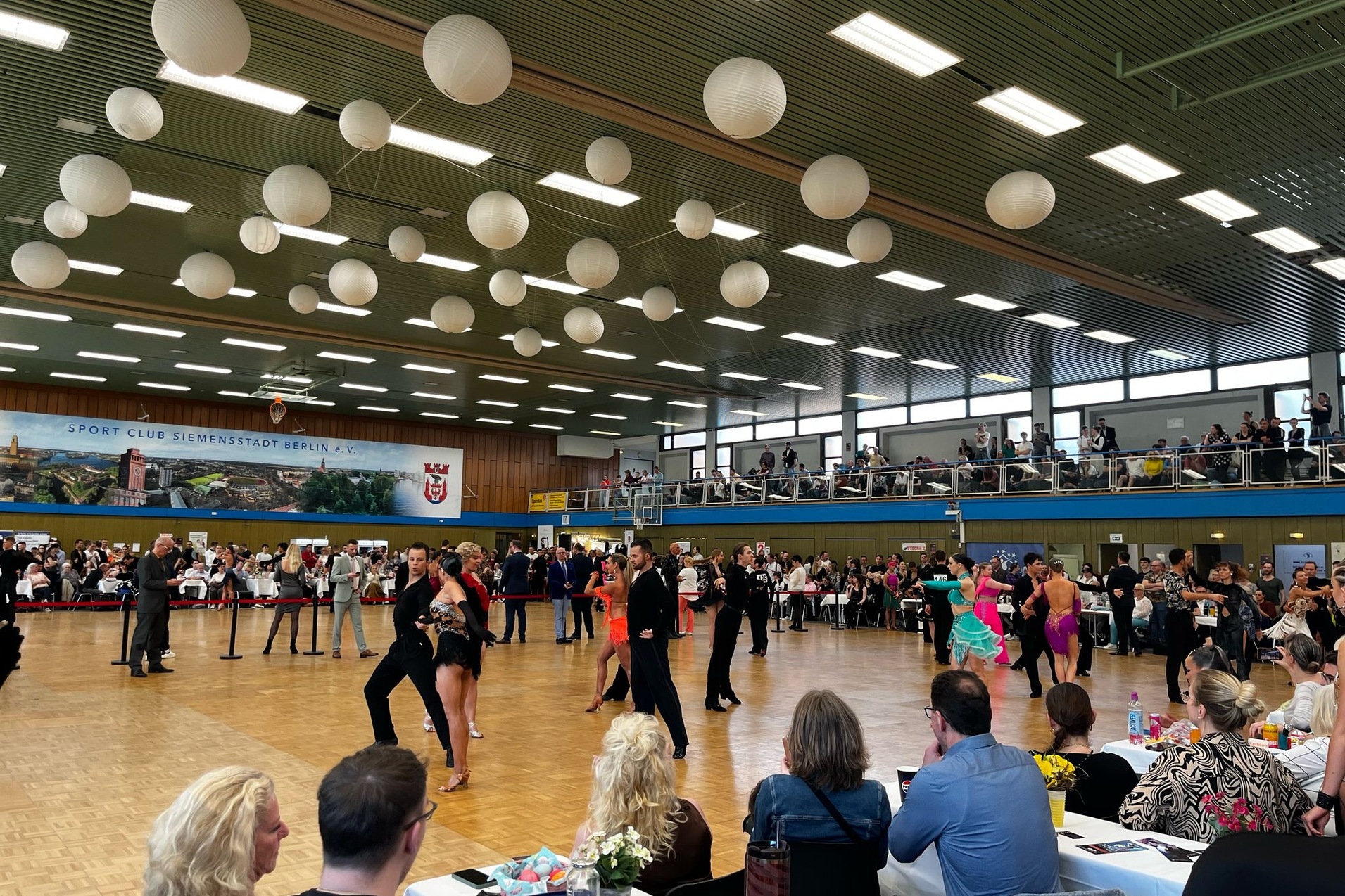 Turnierveranstaltung mit T&auml;nzern in bunter Tanzkleidung, Publikumsbereich und dekorativen Papierlaternen im Hintergrund.