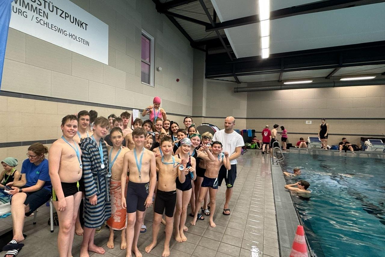 Gruppe von Schwimmern in Badebekleidung mit Medaillen, posieren am Beckenrand eines Hallenbades.