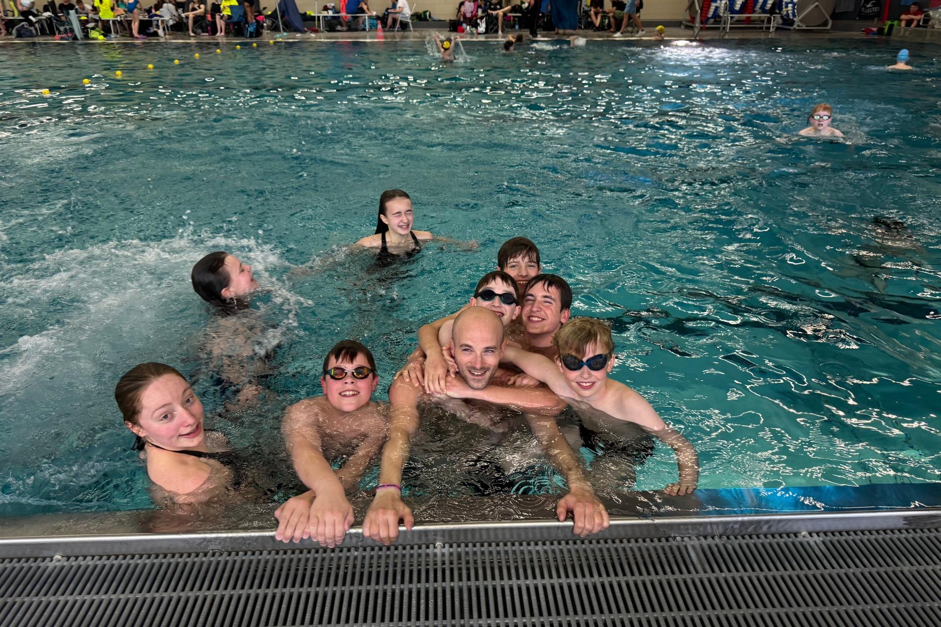 Gruppe von Kindern und einem Erwachsenen im Wasser eines Schwimmbeckens, lächelnd und posierend, mit Schwimmbrillen.