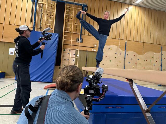 Eine Person balanciert auf einem Schwebebalken, während zwei Kameraleute filmen. Hintergrund mit Kletterwand und Matten.