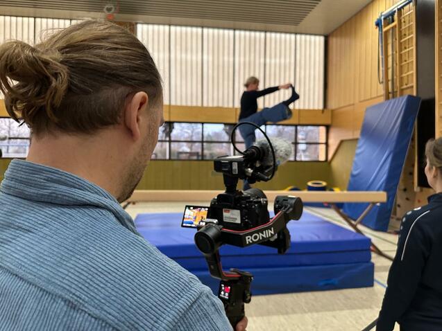 Eine Person filmt mit einer Kamera, während eine andere Person eine Übung an einem Schwebebalken demonstriert.