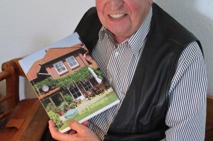 Lächelnder älterer Mann hält ein Buch mit einem Hauscover in der Hand, sitzt an einem Tisch mit bestickter Tischdecke.