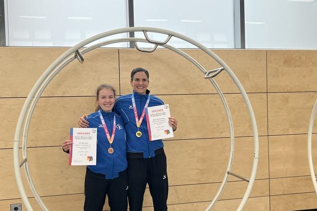 Zwei Athletinnen in blauen Sportjacken stehen in einem großen Holzring und halten Medaillen und Urkunden in den Händen.