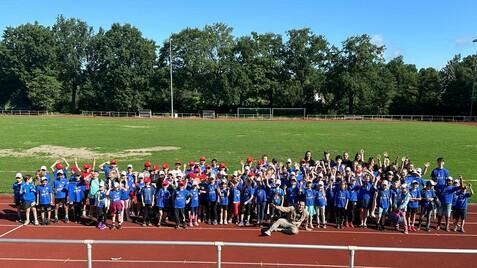 Gruppierung von Kindern in blauen T-Shirts und roten Kappen auf einer Laufbahn, umgeben von Bäumen und einem grünen Feld.